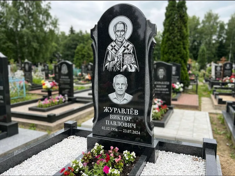 Памятник с гравировкой иконы: стела с образом в полный рост на обороте — гравировка образа Матроны Московской, низкие кусты спиреи в цвету, работа «Лебедева» в Перми